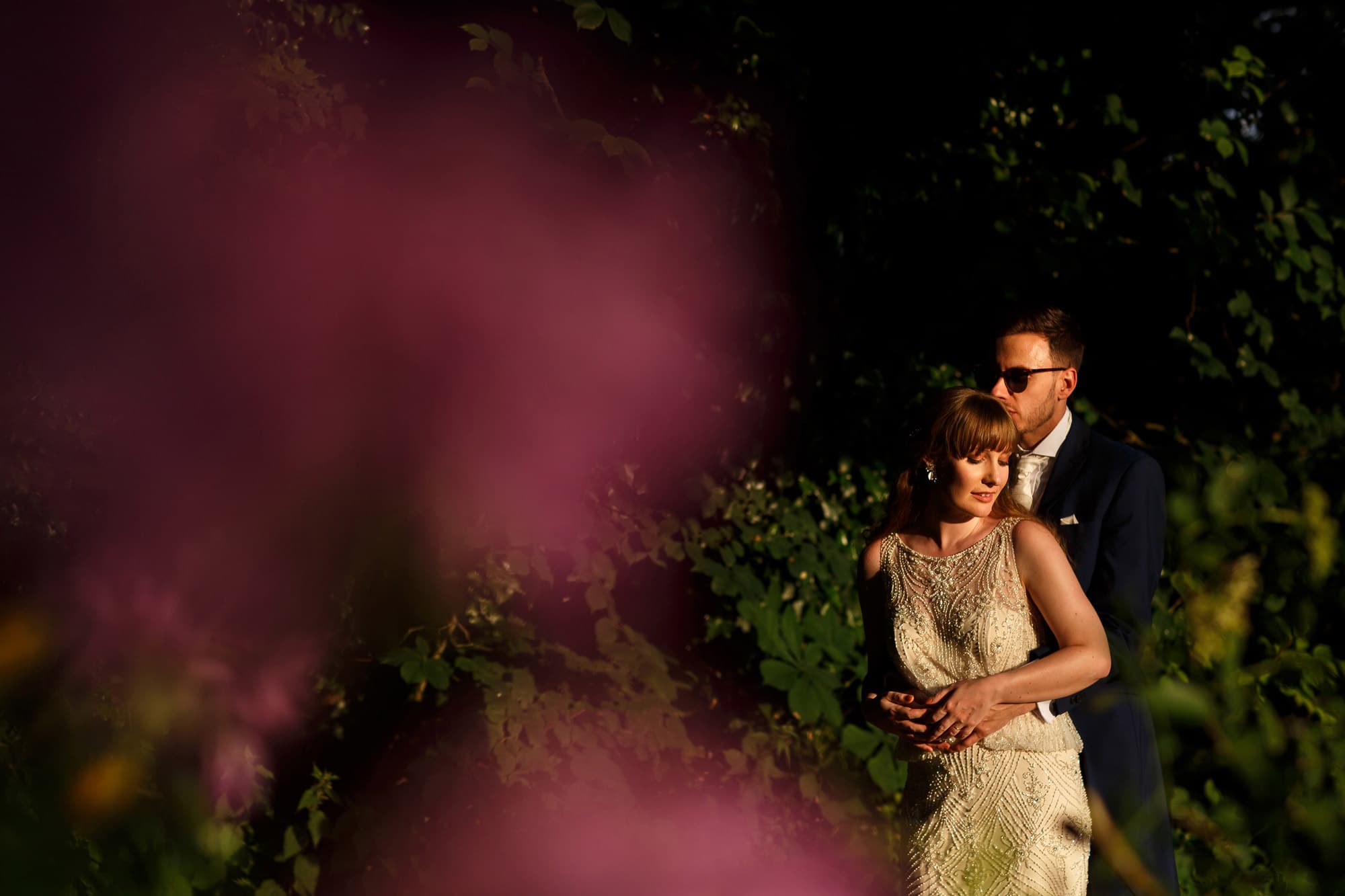 Sexy bride and groom portrait in direct sunlight. Bride wearing beaded wedding dress and groom wearing navy suit and sunglasses.