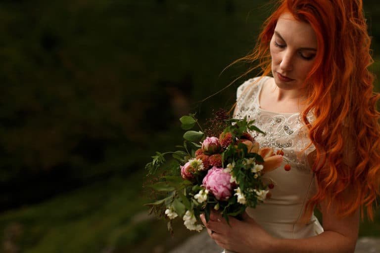 Curly red head bride looking down into colourful DIY wedding bouquet as her hair blows in the wind.