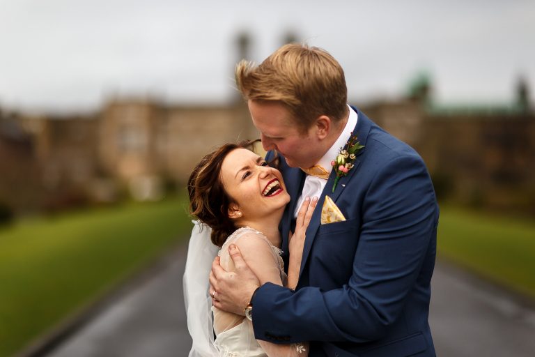 Bride with brunette hair and dark lipstick laughing in her grooms arms outside Stonyhurst College.