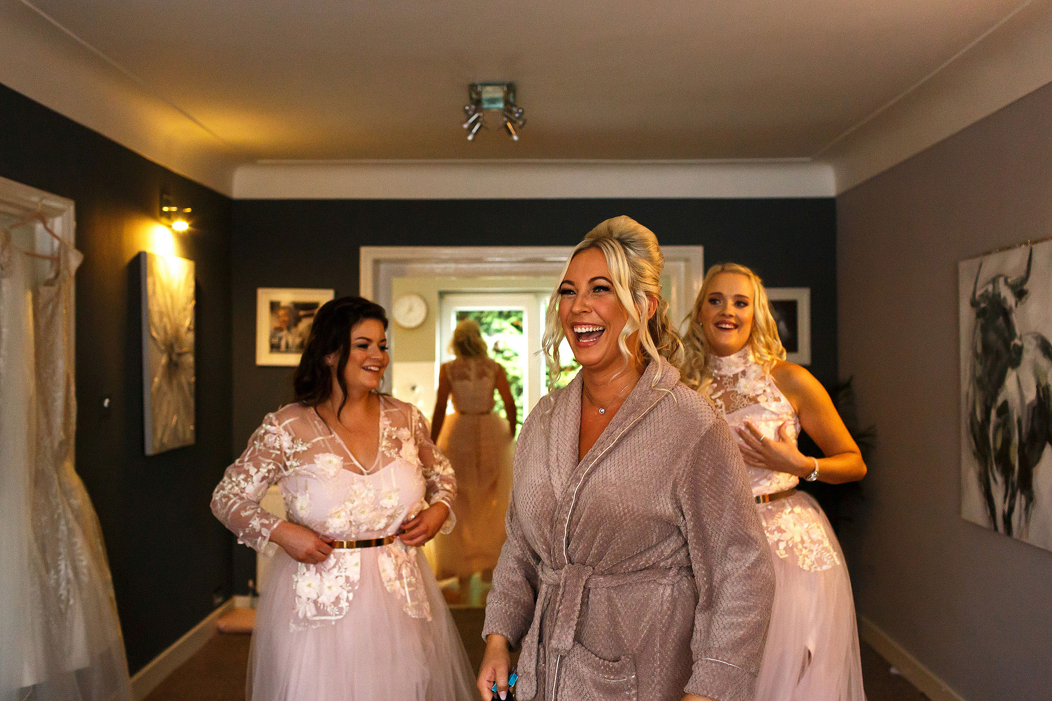 Bride with blonde hair laughing as she looks out the window with bridesmaids behind her