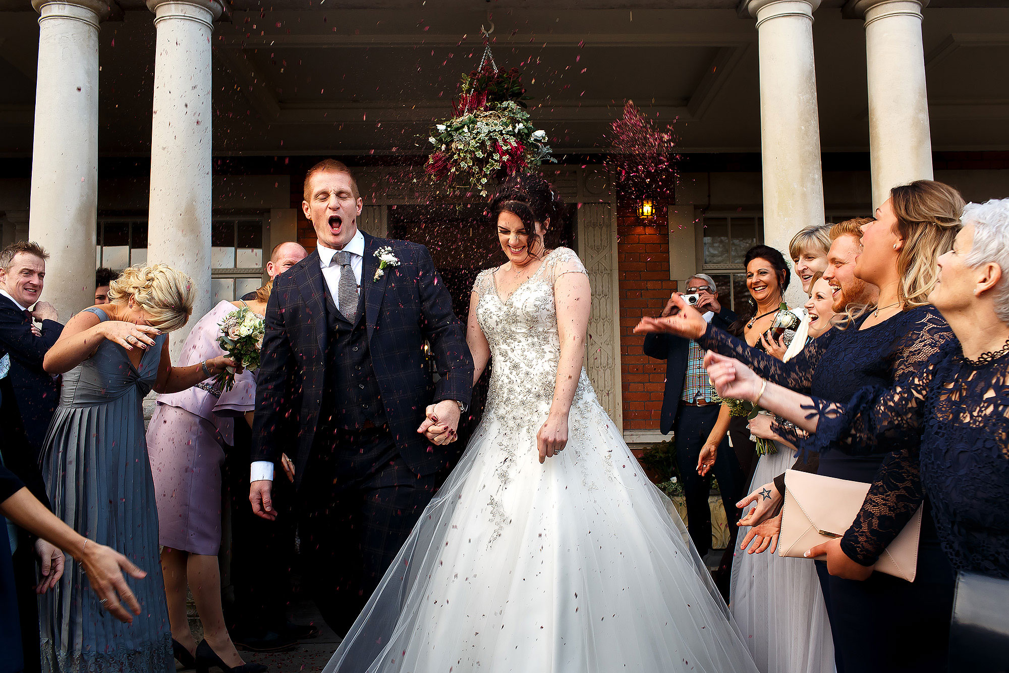 Bride and groom walking out of the pillared entrance as wedding guests throw deep red confetti | Eaves Hall wedding photography by Toni Darcy