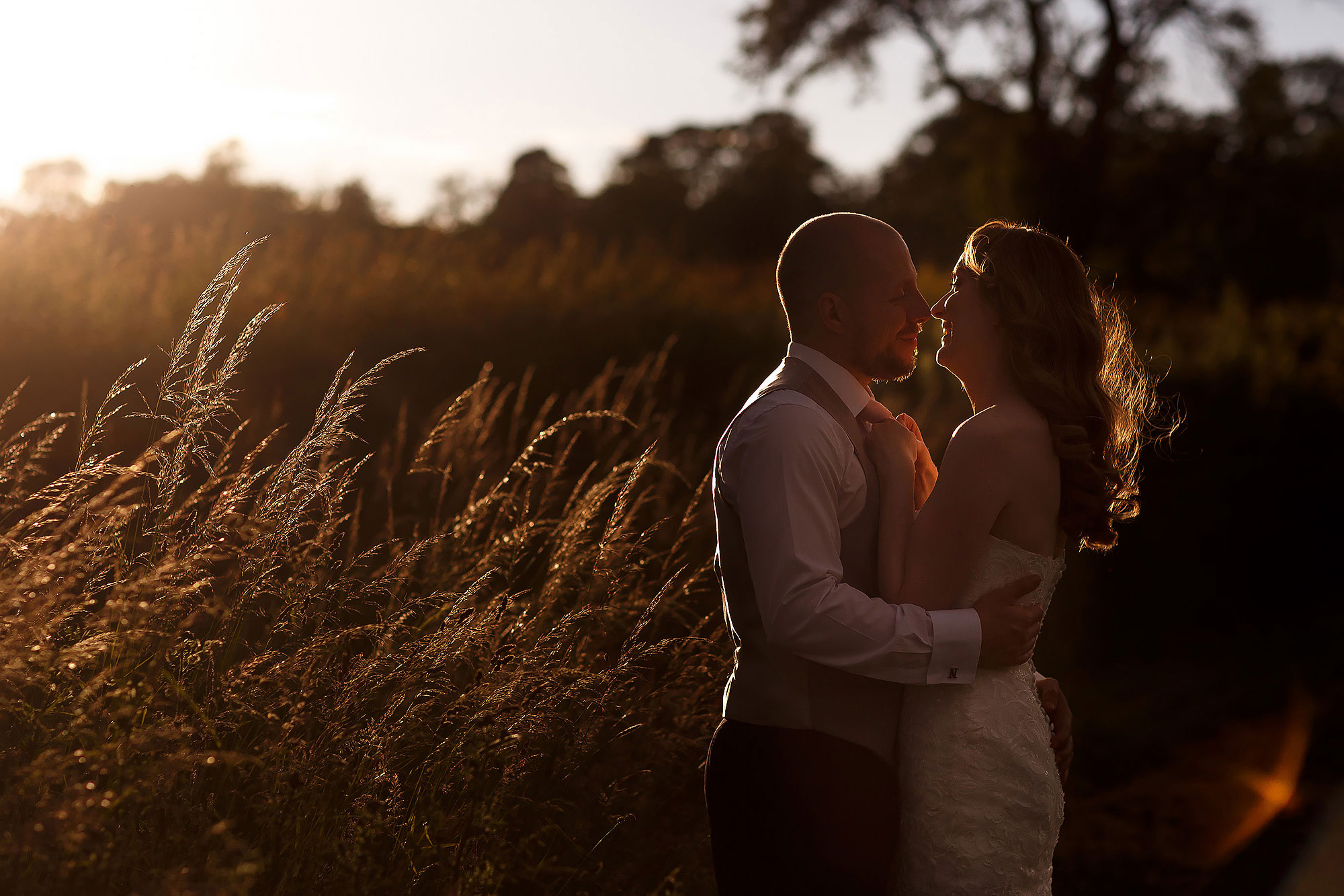 Bride and groom cuddled up in long grass with the sunset behind them