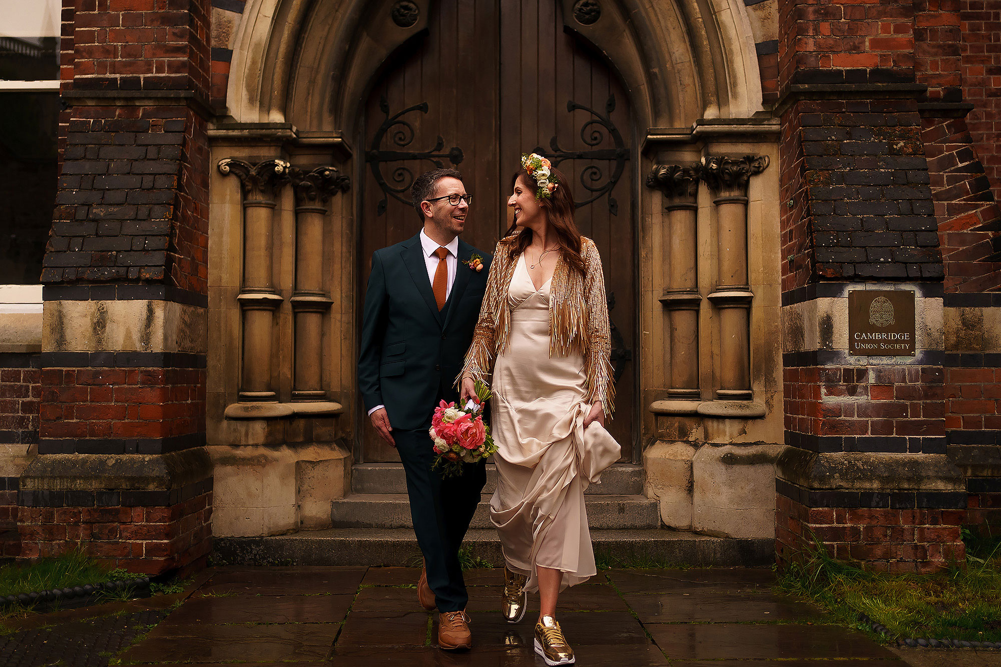 Bride wearing flower crown and cream silk dress and sequin gold jacket with gold trainers holding flowers walking next to groom in blue suit wearing tan nike trainers. Both walking outside Cambridge Union on the footpath.