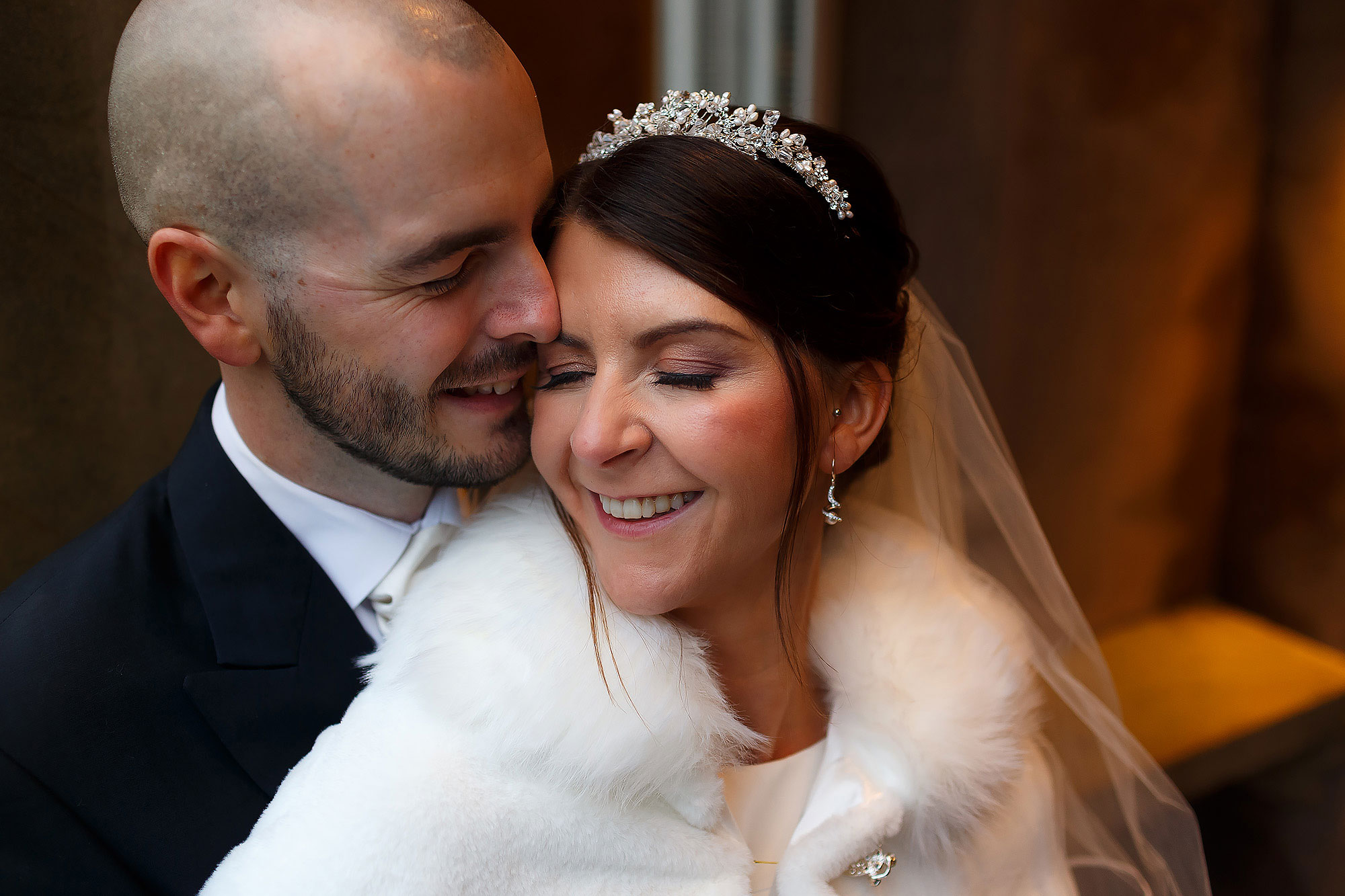 Bride wearing tiara with brown hair and fur shawl. Groom giving kisses on the cheek. In the doorway at Mitton Hall.