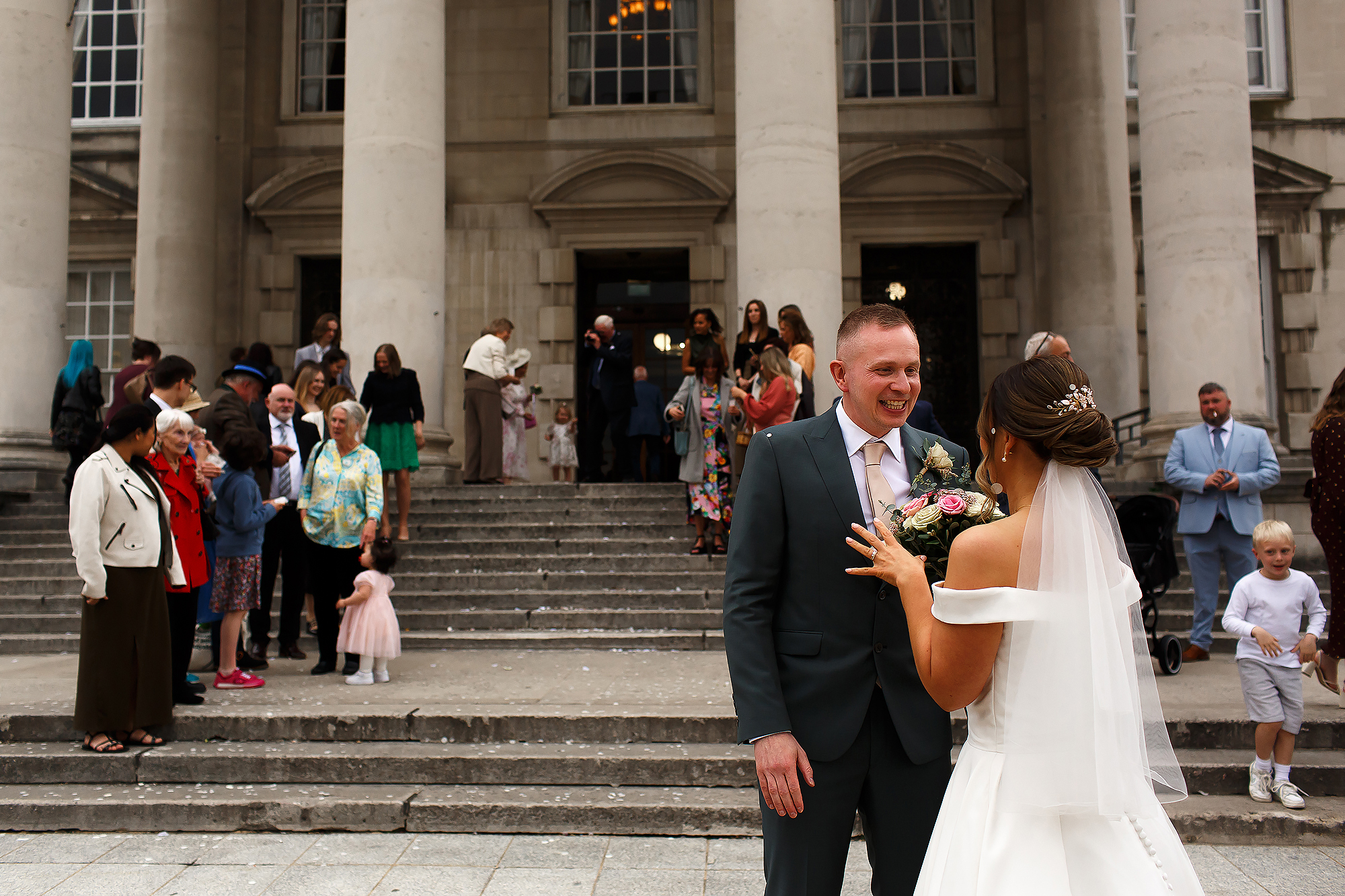 Bride and groom at the bottom of the steps of Leeds Civic Hall with the guests on the steps behind them