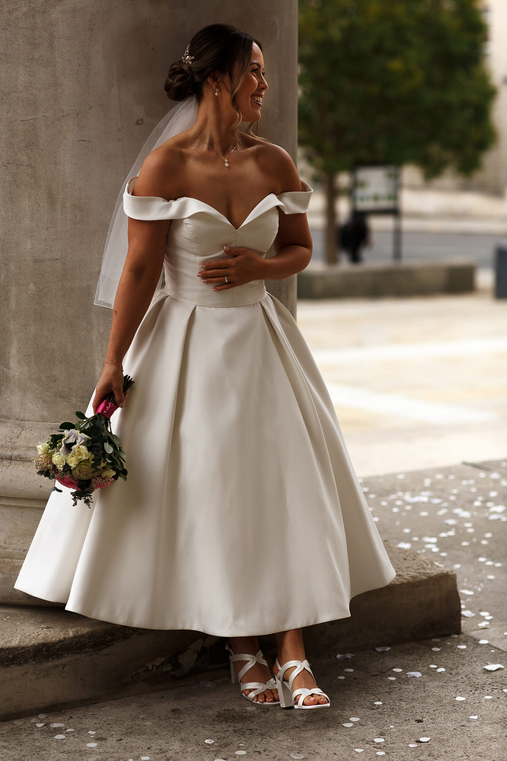 Bride stood by the columns of Leeds Civic Hall wearing tea length wedding dress holding flowers by her side